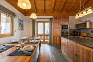 Interior de madera con una mesa de comedor preparada para cuatro personas, una cocina con armarios de madera y vistas a través de una puerta de cristal.