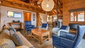 A spacious living area with a wooden ceiling, a large round light fixture, a sofa, two blue leather chairs, a coffee table, and a television. - Chalet Elbaz - Swiss Chalet With Breathtaking Views (Riddes)