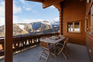 A wooden balcony with a table and chairs, overlooking a mountainous landscape with snow-capped peaks.
