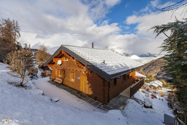 A wooden cabin with a sloped roof, surrounded by snow-covered landscape and mountains in the background.