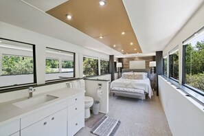 Ein modernes Schlafzimmer mit einem großen Bett, ein Badezimmer mit Waschbecken und Toilette sowie große Fenster mit Blick auf Grünflächen.