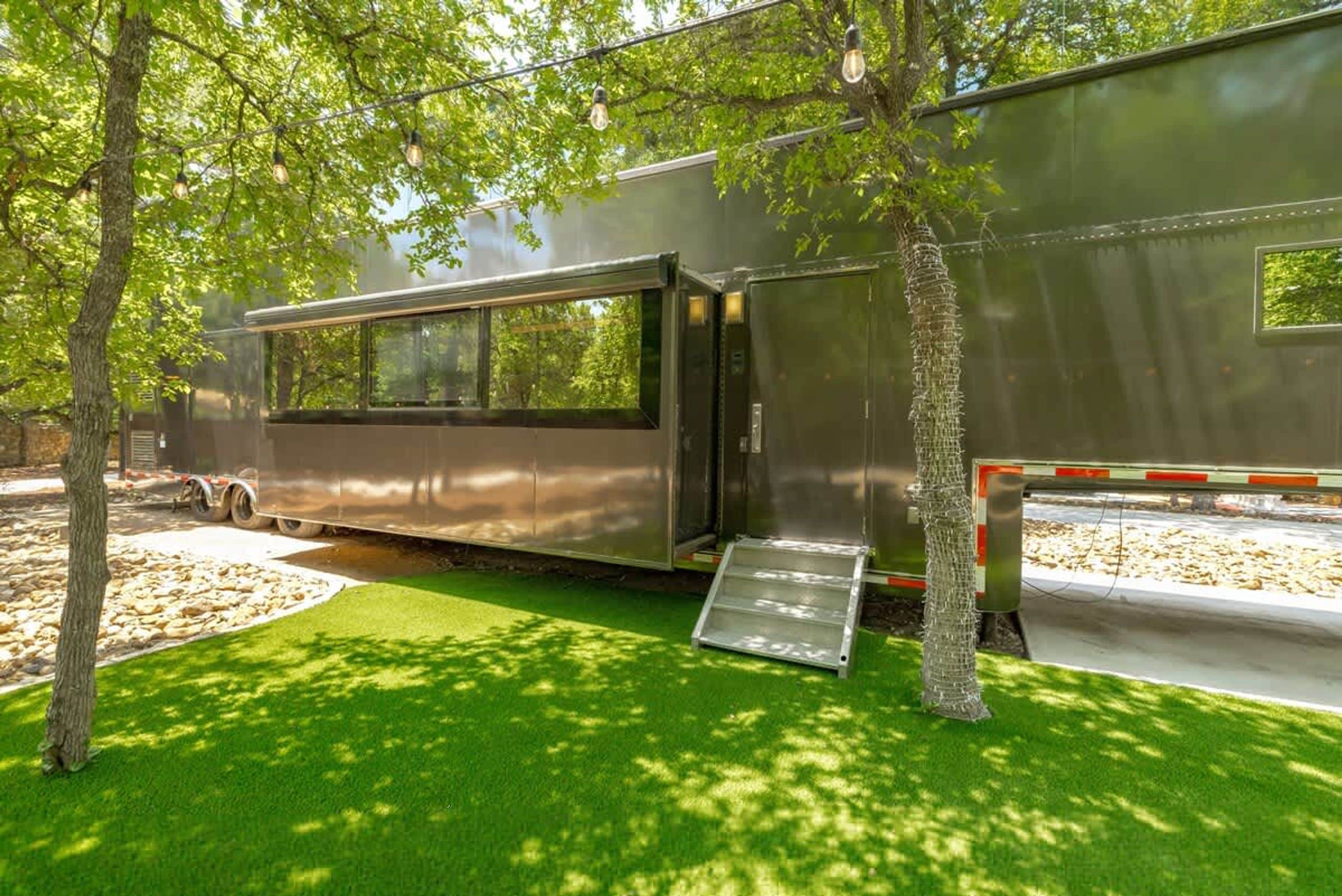 Un camping-car moderne doté d’une porte coulissante, d’un petit espace extérieur avec du gazon synthétique et d’un arbre à proximité.