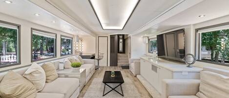 Una sala de estar moderna con sofá, mesa de centro y televisor. Hay ventanas grandes y una escalera que sube a otro nivel.