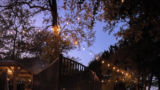 A stone pathway leading to a wooden deck with string lights, surrounded by trees and a stone wall. - Studio 7744 Ranch Austin Venue and Rentals (Austin)