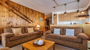 A cozy living room with two brown sofas, a wooden wall with skis, a wooden coffee table, and a kitchen area with wooden cabinets and pendant lights. - 6 Person ski in out Apartment Great Views Garage (Riddes)