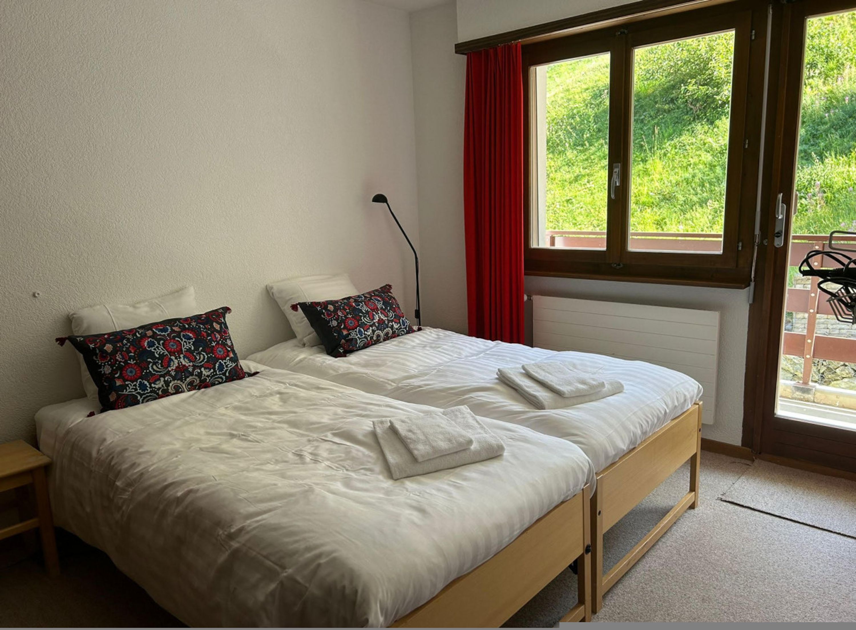 A double bed with white bedding and two decorative pillows, a bedside table, a window with red curtains, and a view of greenery outside.