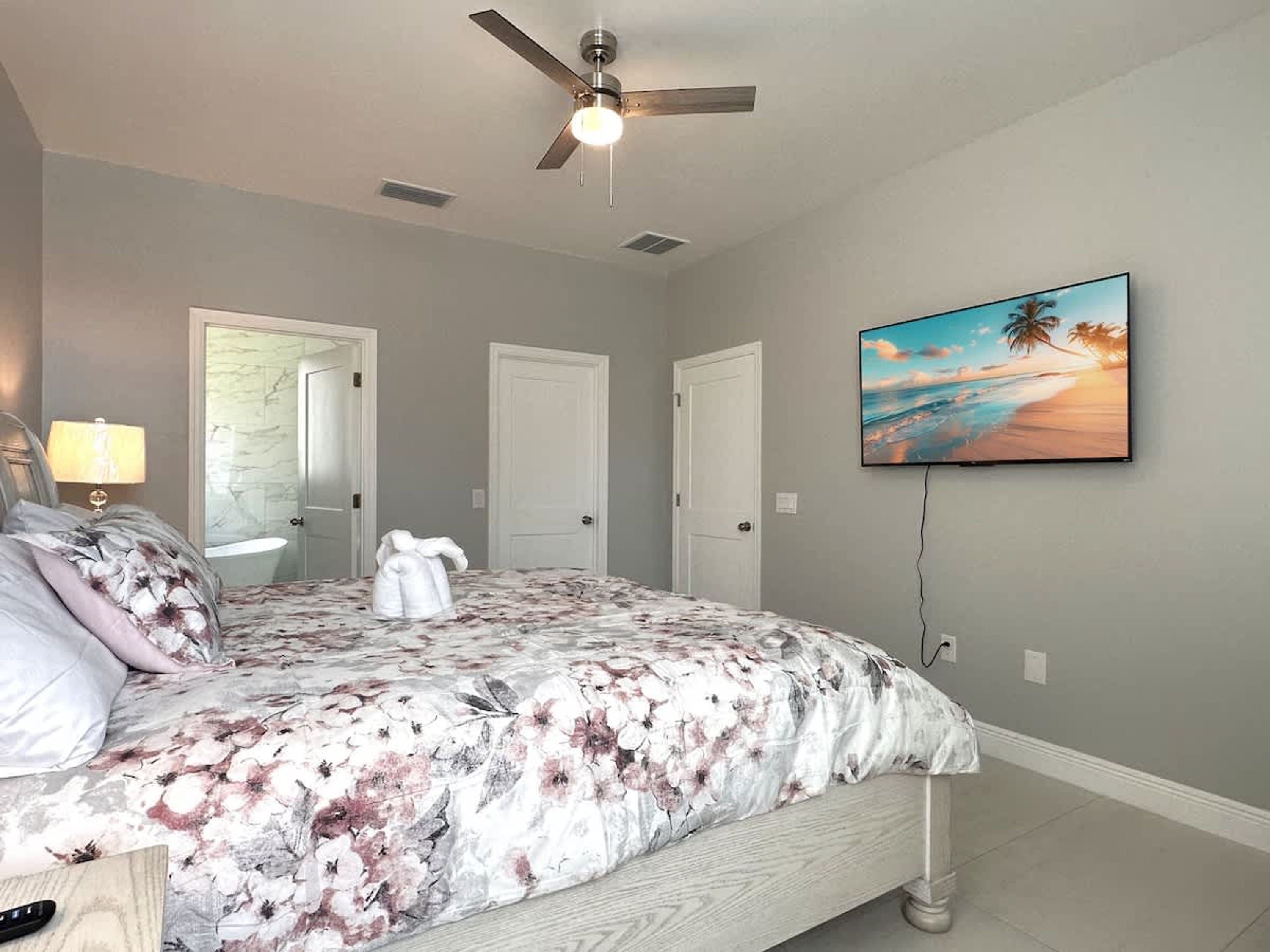 Une chambre à coucher avec un lit, un ventilateur de plafond, un tableau représentant une scène de plage et une porte.