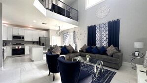 A spacious living area with a grey sofa, blue armchairs, and a glass coffee table. A kitchen with white cabinets and stainless steel appliances is visible in the background.