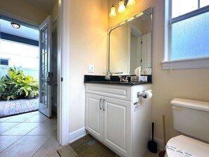 Une salle de bain avec un meuble-lavabo blanc, un grand miroir et des toilettes. Il y a une fenêtre avec du verre dépoli et une porte qui donne sur un patio agrémenté de plantes.
