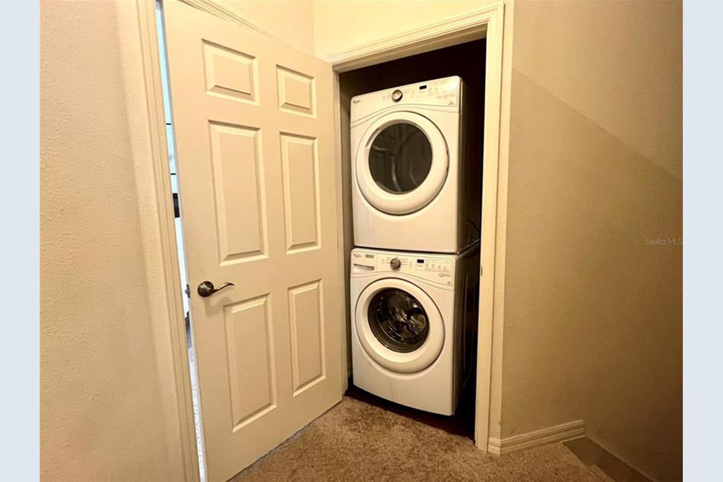 A laundry room with a front-loading washer and dryer, beige walls, and a carpeted floor.