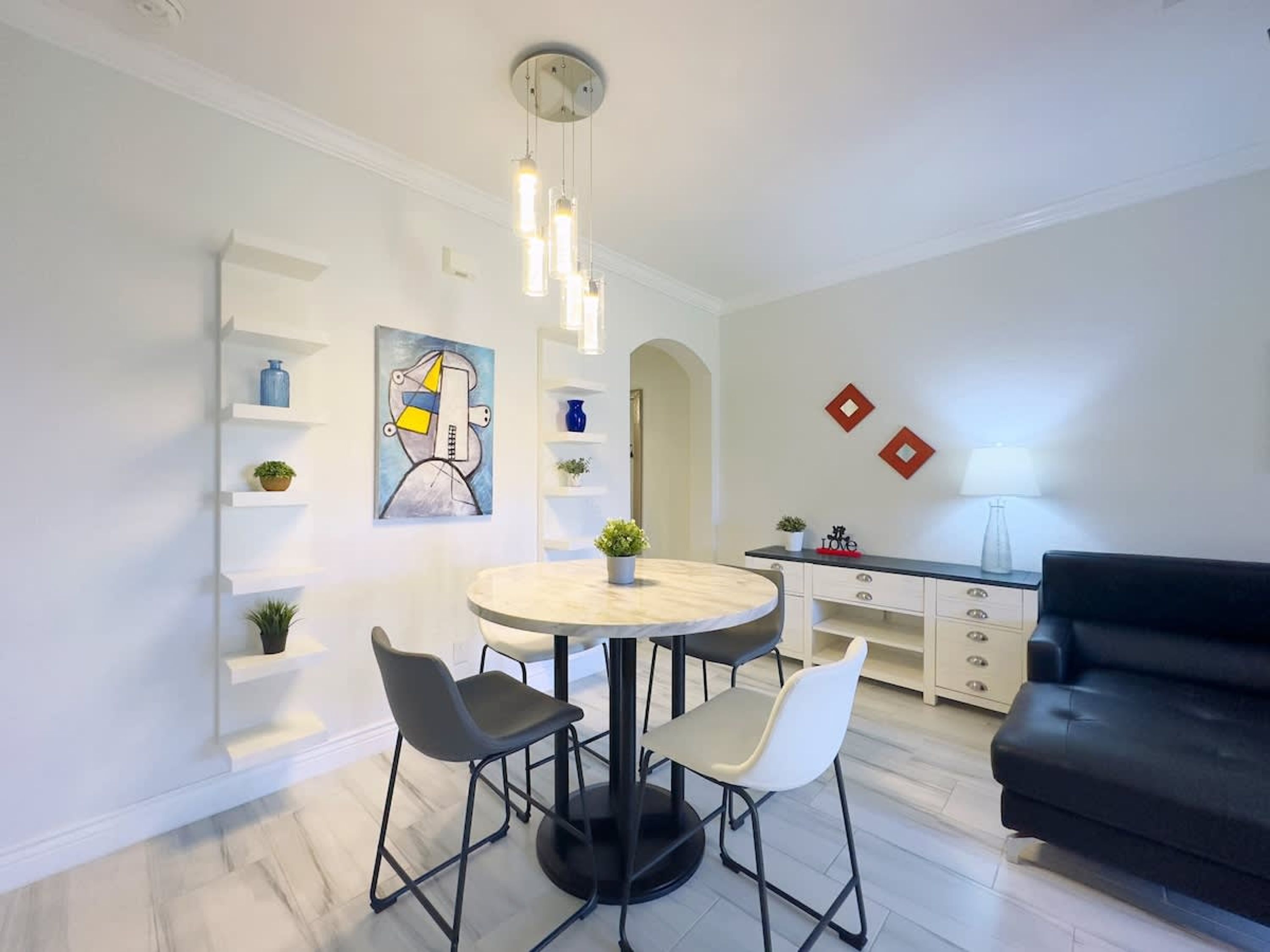 Un soggiorno moderno con un tavolo da pranzo rotondo, due sedie, un divano nero e un tavolino. C'è un quadro appeso alla parete e una lampada su un comodino.