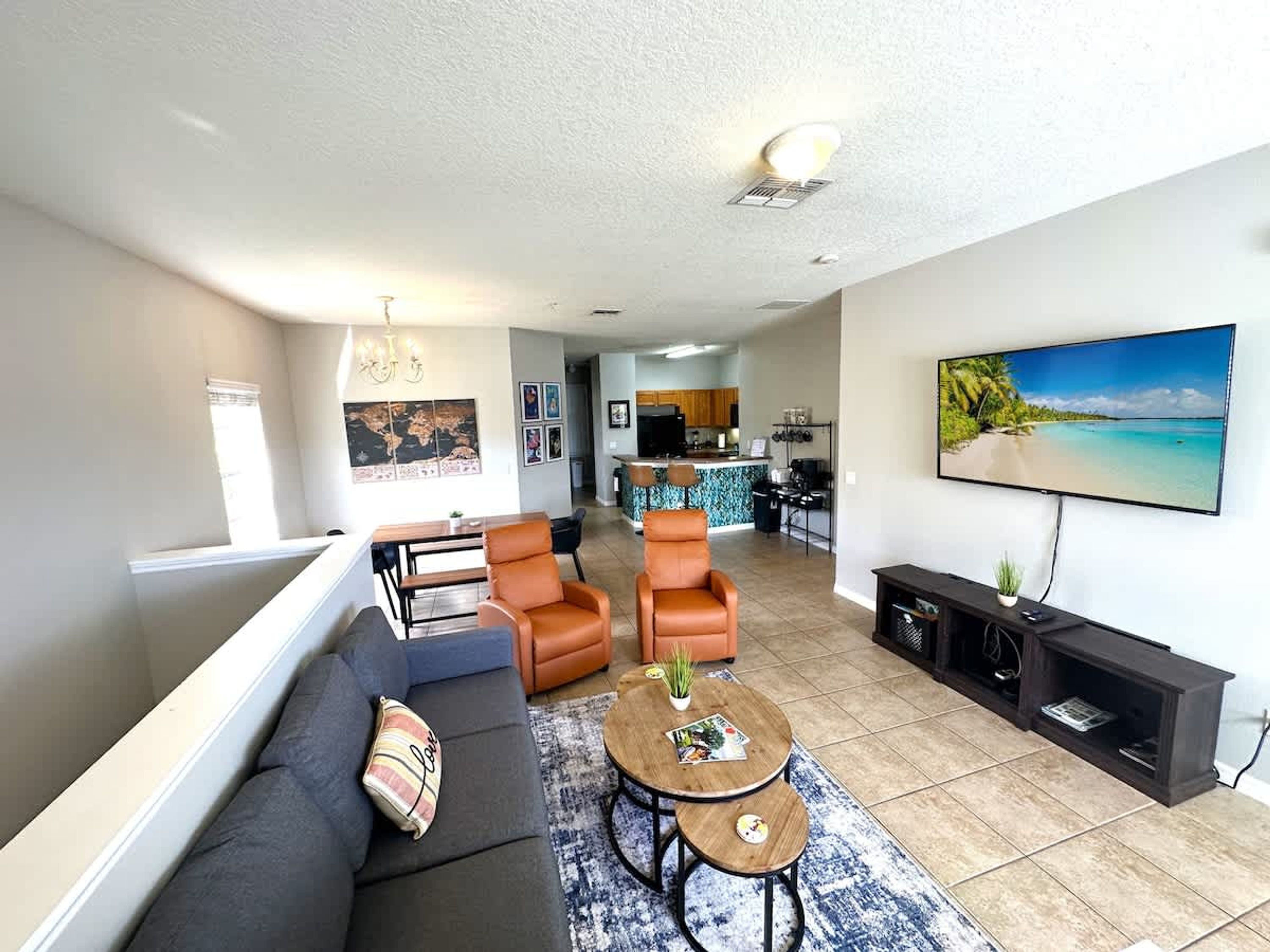 A modern living room with a sofa, two armchairs, a coffee table, and a TV. There is a kitchen in the background.