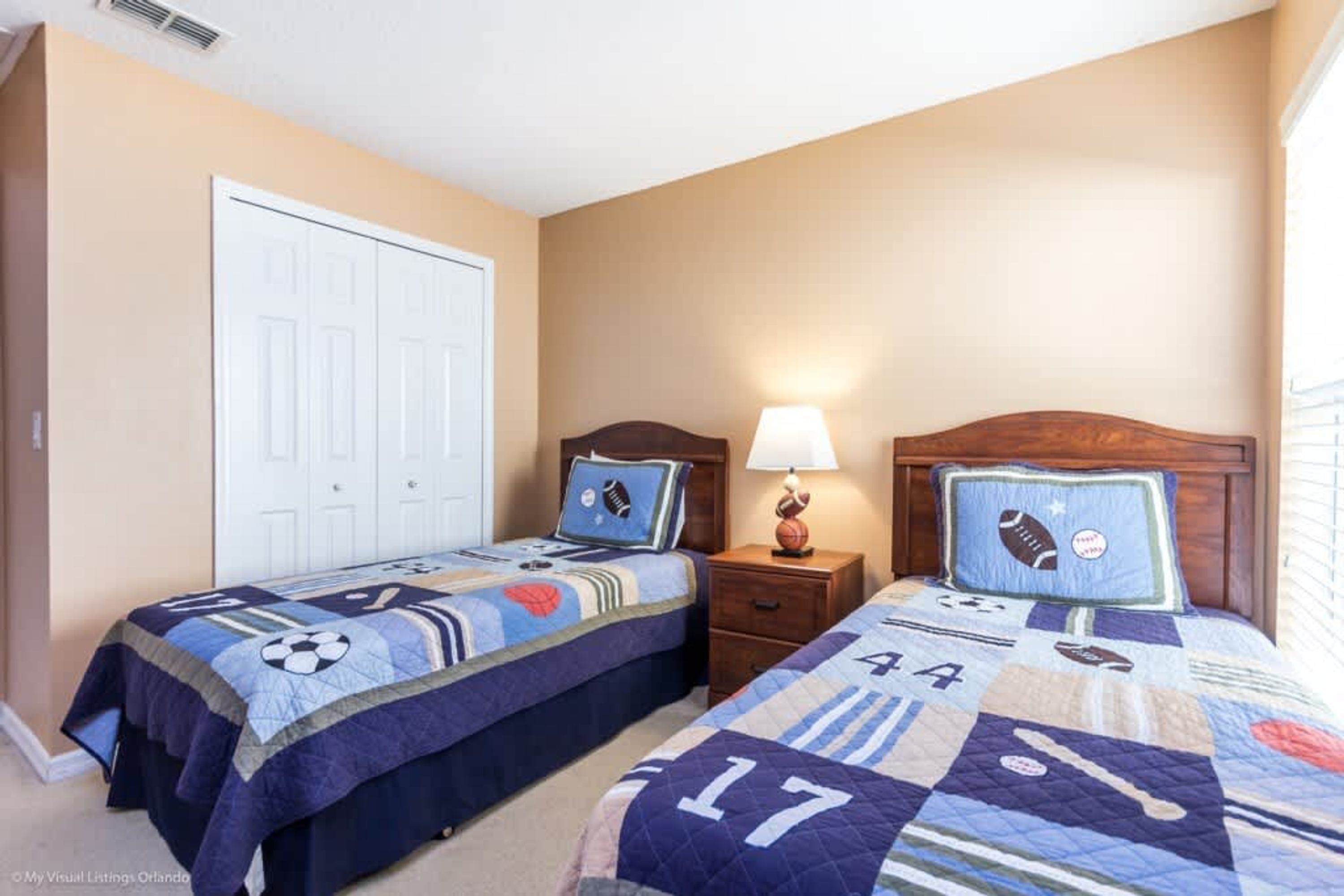 A bedroom with two beds, each with a sports-themed quilt. There is a white door and a nightstand with a lamp.
