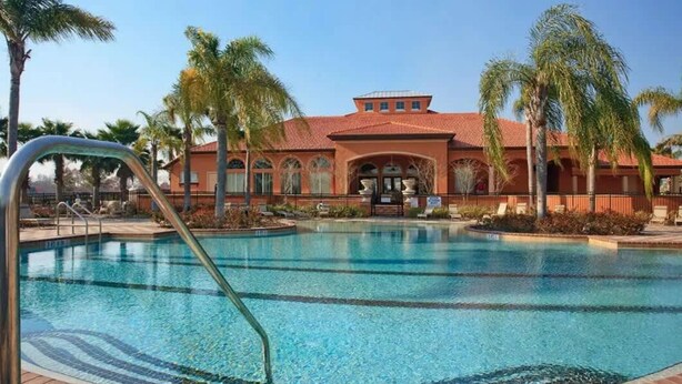 A swimming pool with a clear blue water, surrounded by palm trees and a building with arched windows and a red roof.