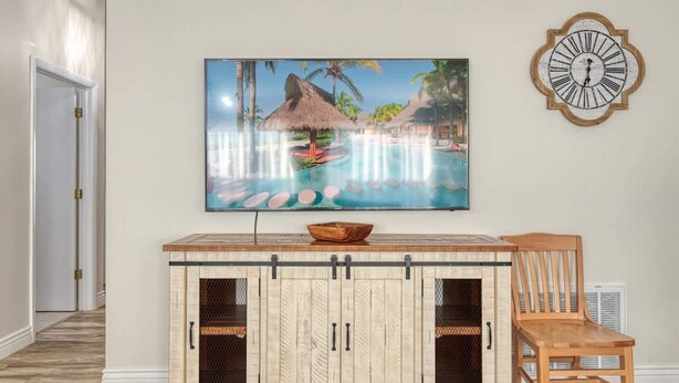 Ein Zimmer mit Holzschrank, Esstisch, Stuhl, einem Bild eines tropischen Resorts und einer Wanduhr.