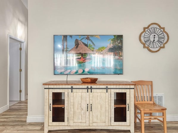 Een kamer met een houten kast, een eettafel, een stoel, een schilderij van een tropisch resort en een wandklok.