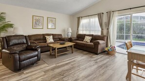 Ein Wohnzimmer mit Deckenventilator, zwei Sofas, einem Couchtisch, einer Lampe und Blick auf einen Garten durch ein großes Fenster.