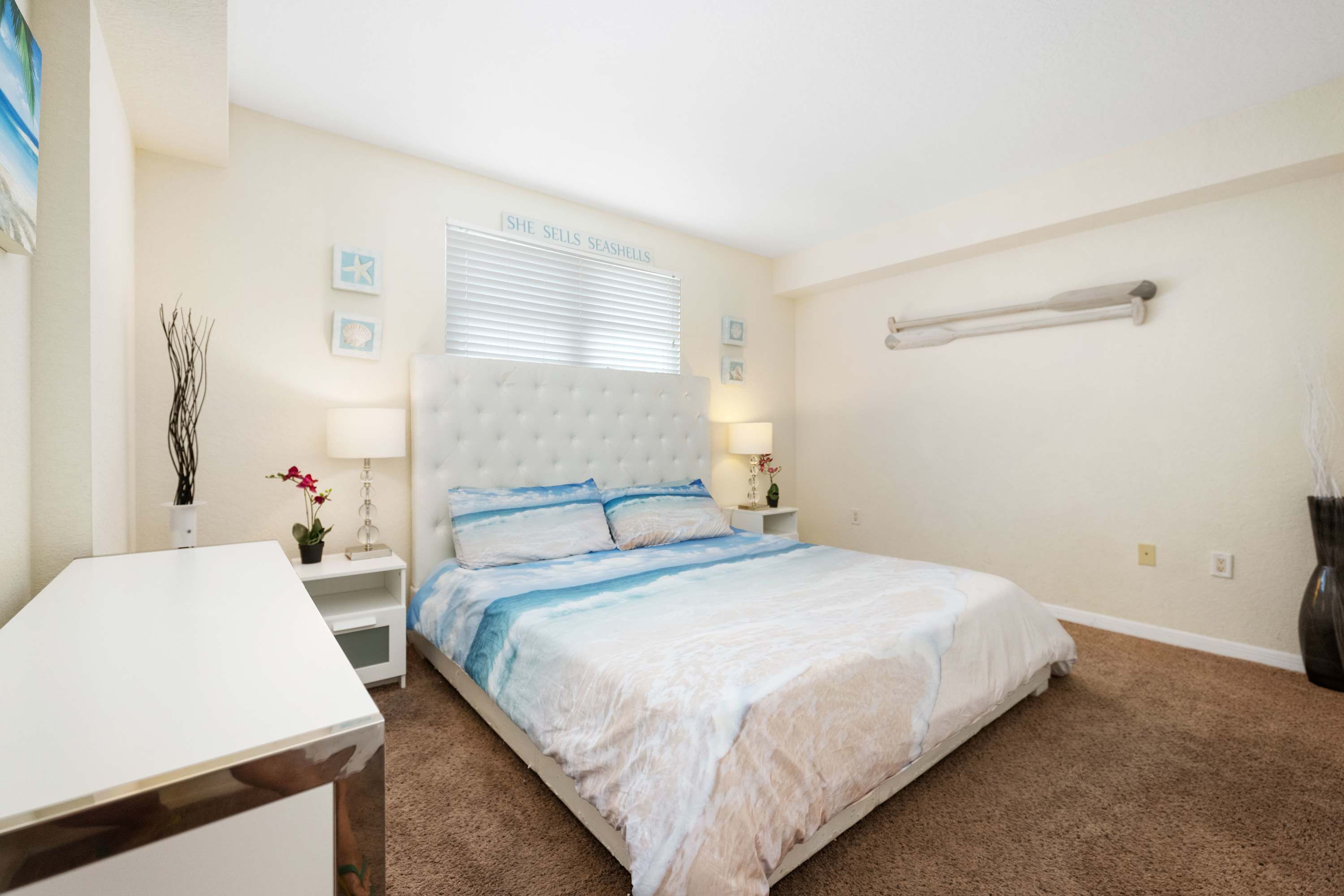 A bedroom with a large bed, a bedside table, a nightstand, a wall-mounted shelf, and a window with blinds.