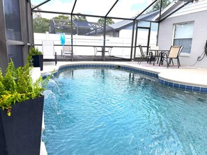Piscina con cerramiento de vidrio, rodeada de mobiliario exterior y plantas en macetas.