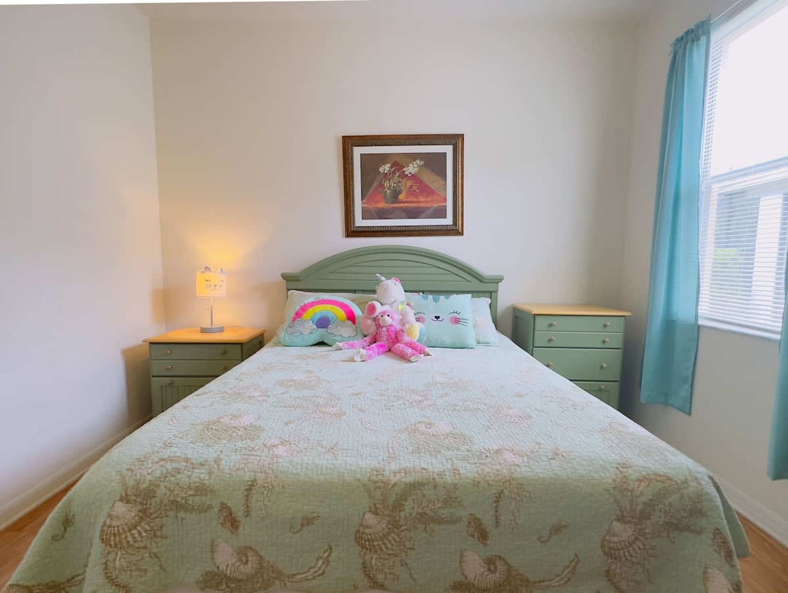 A bedroom with a bed, a nightstand, a lamp, a framed picture on the wall, and a window with curtains.