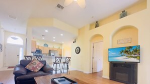 A living room with a sofa, coffee table, and TV. There is a kitchen in the background with a dining area. - 4Bd Pool Villa in Calabay Parc Close to Disney Sleeps 8 (Haines City)