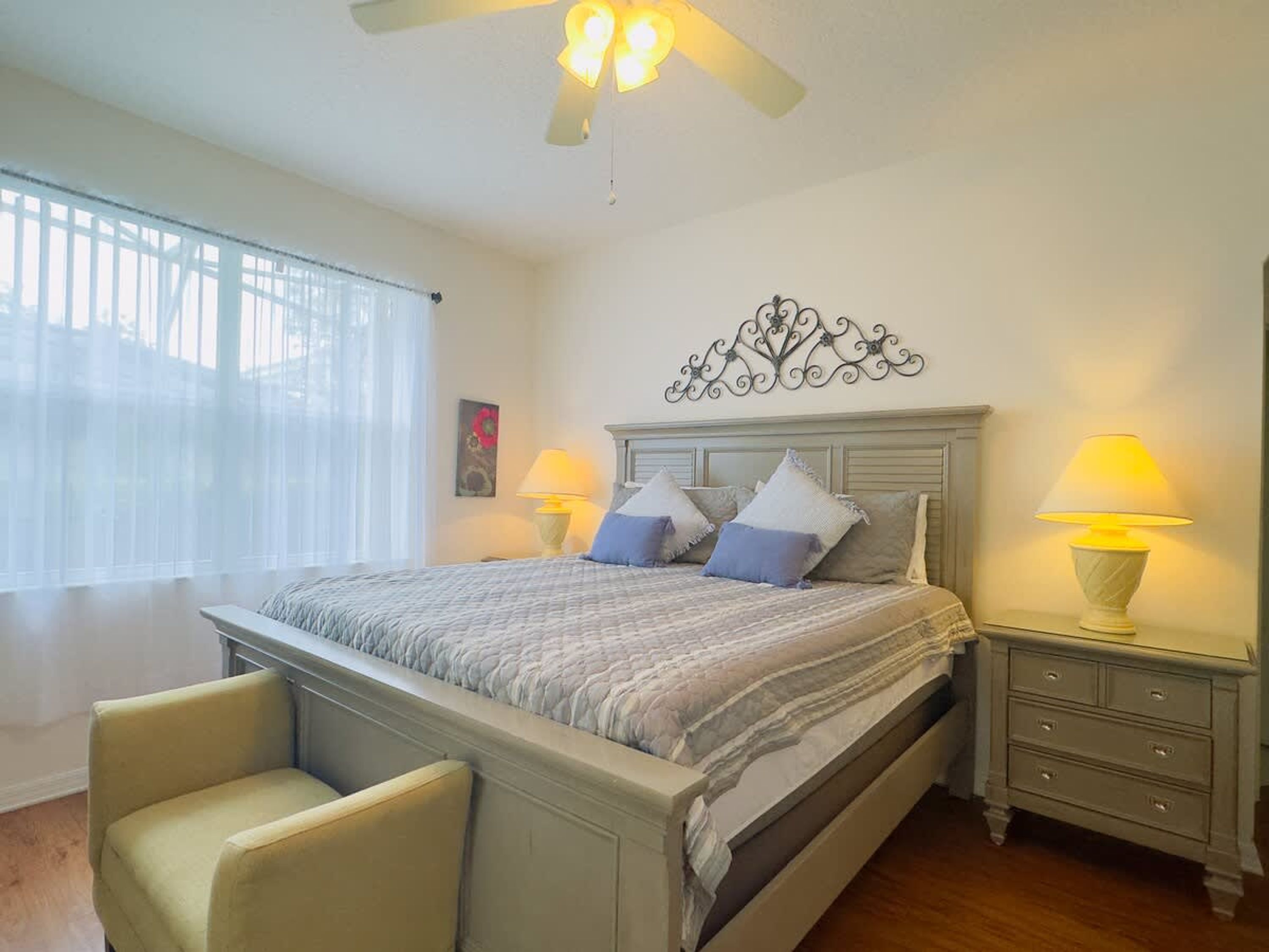 A bedroom with a bed, two bedside tables with lamps, a chair, a ceiling fan, and a window with blinds.