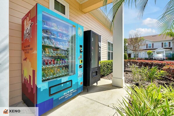 A colorful vending machine with a variety of drinks and snacks, located outside a building with a covered entrance.