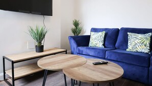 A living room with a blue sofa, two round wooden tables, a flat-screen TV mounted on the wall, and two potted plants. - Charming 2 Bed-ensuites Fast Wi-fi (Coventry)