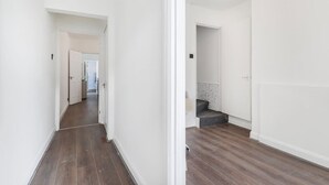 A hallway with wooden flooring and white walls, leading to a room with a grey sofa and a small table. - Charming 2 Bed-ensuites Fast Wi-fi (Coventry)