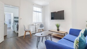 A modern living room with a blue sofa, a small table, and a TV mounted on the wall. - Charming 2 Bed-ensuites Fast Wi-fi (Coventry)