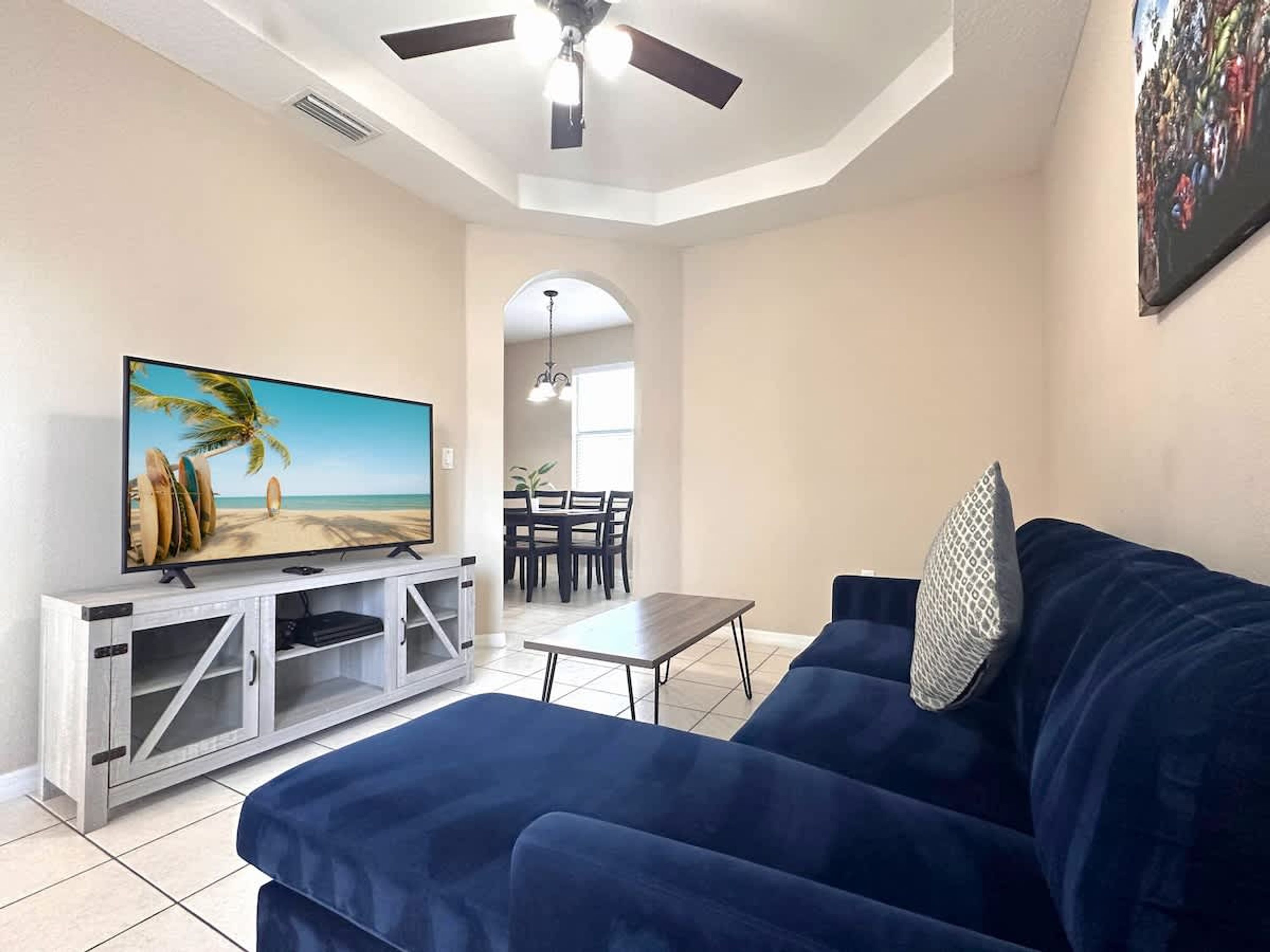 Een woonkamer met een televisie op een witte tv-meubel, een blauwe bank, een eethoek met een tafel en stoelen, en een plafondventilator.
