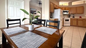 Un coin repas avec une table en bois, quatre chaises et une plante en pot. On aperçoit, au fond, une cuisine avec des meubles et des appareils électroménagers en bois.