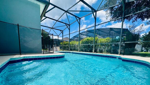 Une piscine d’un bleu éclatant, entourée d’une paroi de verre, bordée de verdure et surplombée par un ciel dégagé.