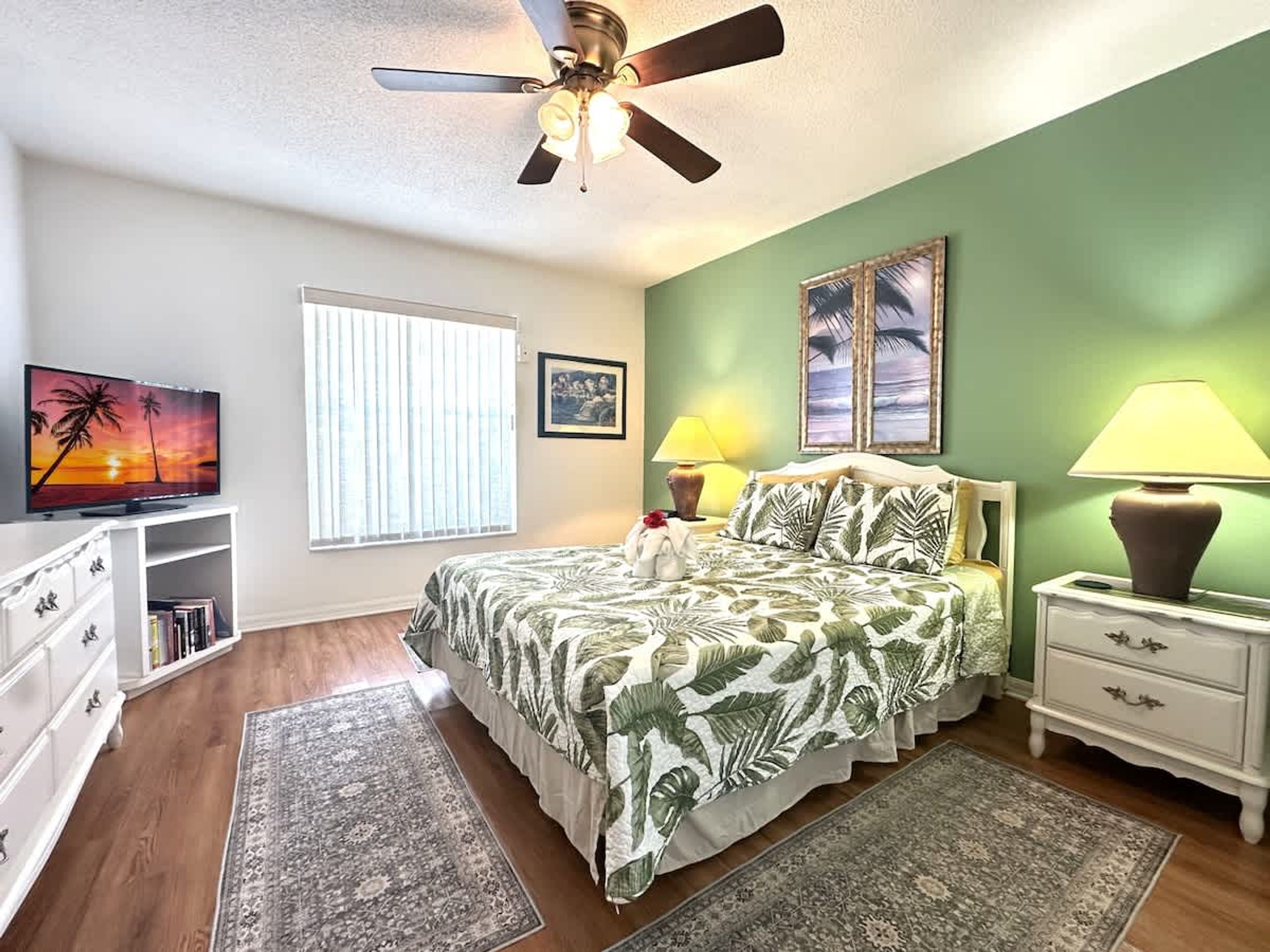 A bedroom with a bed, nightstands, a TV, a ceiling fan, and a window with blinds.