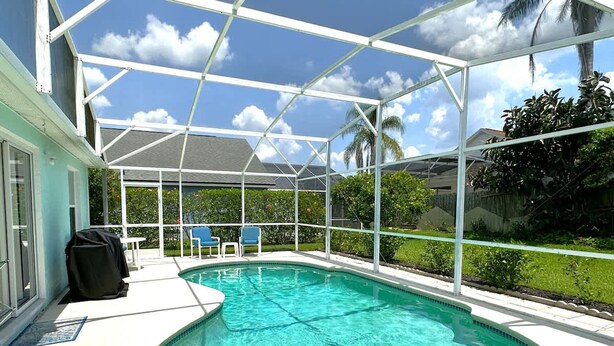 A covered outdoor swimming pool with a glass enclosure, surrounded by greenery and a clear sky.