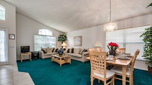 A spacious living room with a teal carpet, a sofa set, a dining table with chairs, and a television. - Villa Near Disney 4 Bd 3 Bt With Private Pool Quiet Comminuty (Davenport)