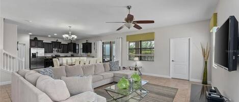 A modern living room with a sofa, coffee table, and TV. There is a kitchen in the background with cabinets and a dining area.