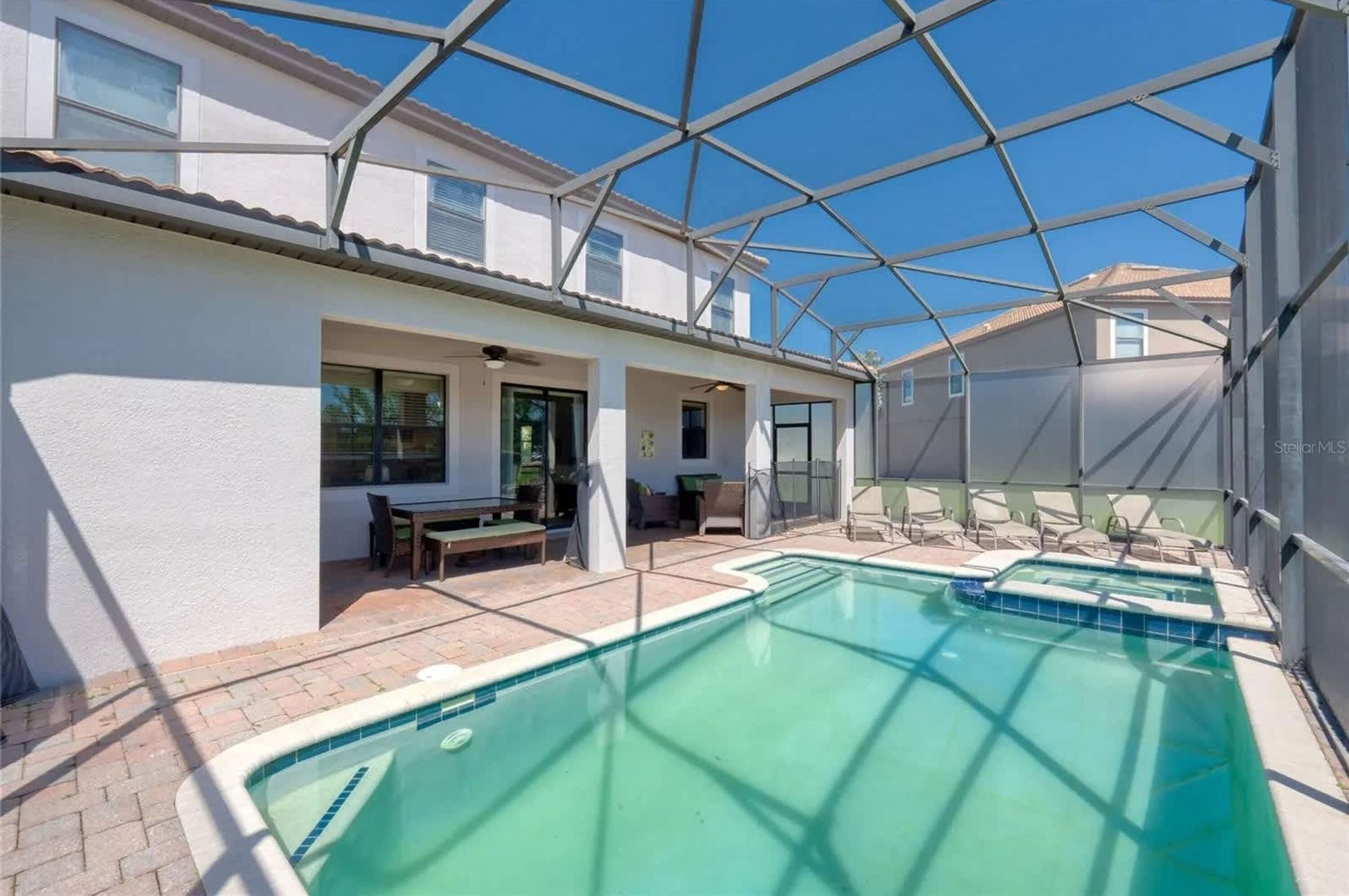 A pool area with a glass-enclosed patio, outdoor seating, and a clear blue sky.