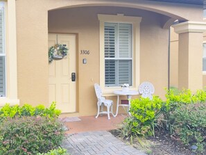 A beige building with a number 3306 on the door, a white chair, a small table, and a window with shutters.
