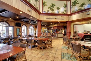 Un restaurante espacioso con techos altos, ventanas amplias y un mural tropical. Mesas y sillas dispuestas para comer.