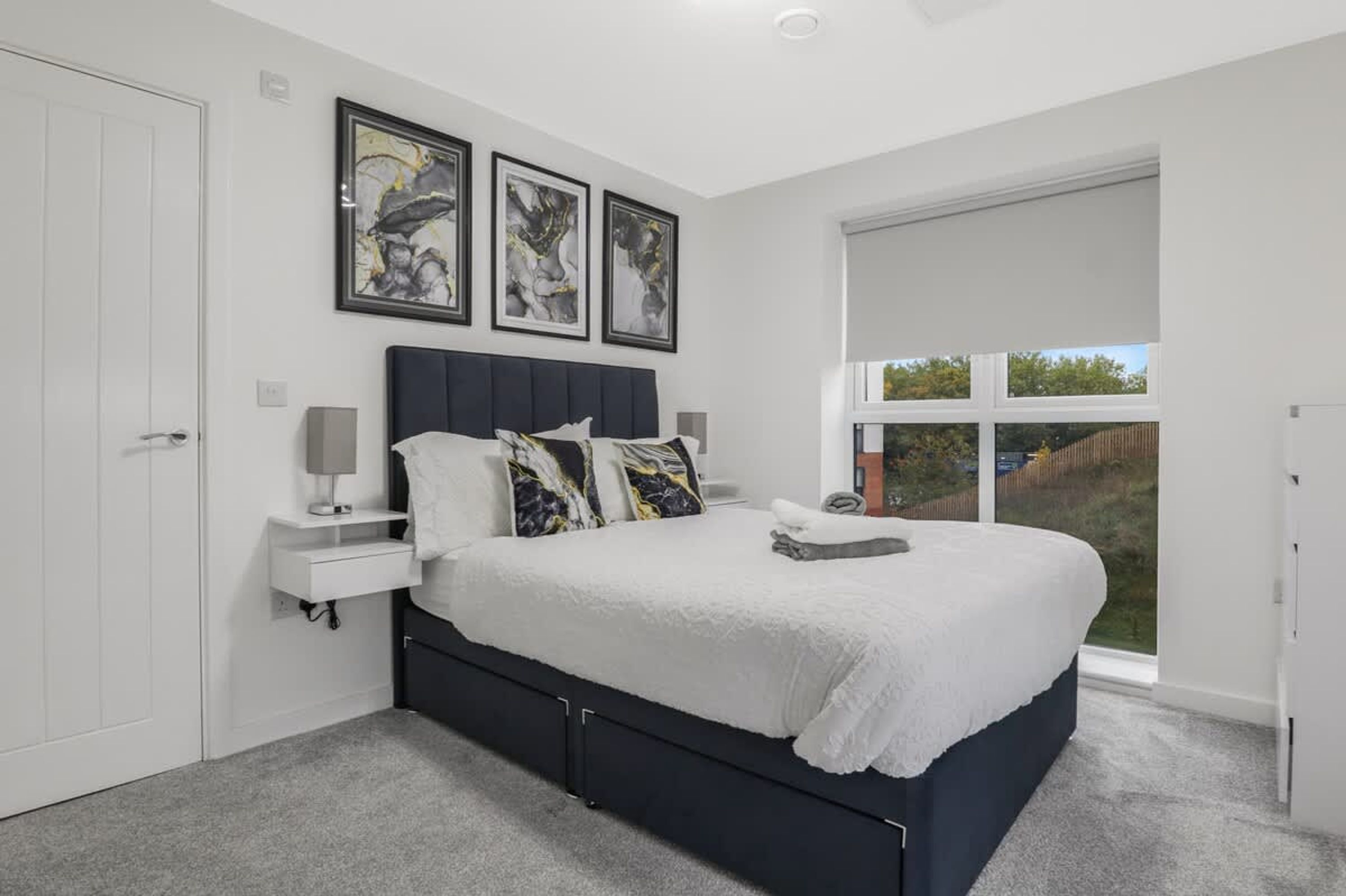 Une chambre avec un grand lit, une porte blanche, une fenêtre donnant sur un beau paysage et trois œuvres d’art encadrées accrochées au mur.