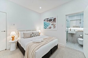 A modern bedroom with a large bed, bedside lamps, a framed picture, and a bathroom with a glass shower. - Coconut Apartment at Palm View Collective (Sydney)