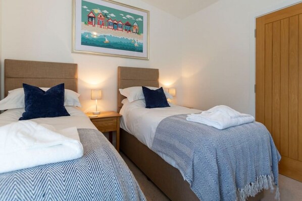 A hotel room with two beds, each with a headboard, bedside tables, and lamps. There is a wooden door and a framed picture on the wall. - The Grove in Cornwall (Bude)