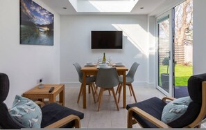 Comedor con una mesa lista para cuatro, un televisor de pantalla plana montado encima y vista al jardín a través de un ventanal corredero.