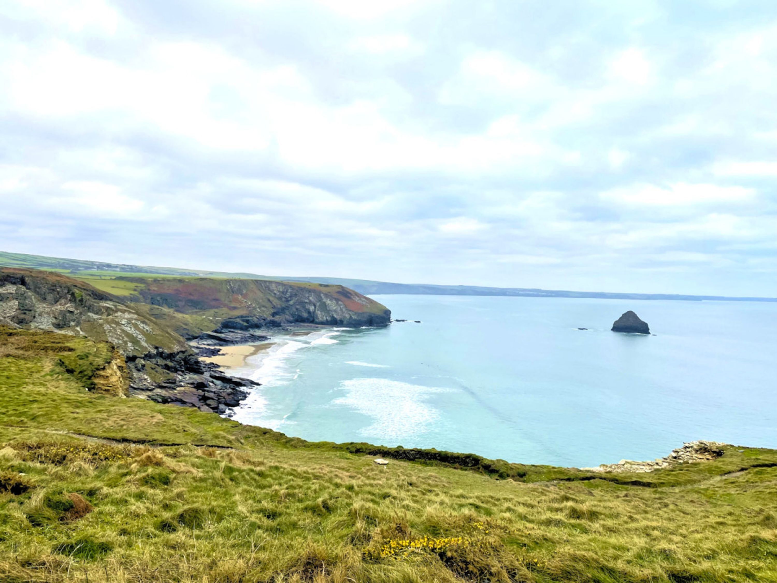A coastal landscape with grassy cliffs, a sandy beach, and a large rock formation in the sea.