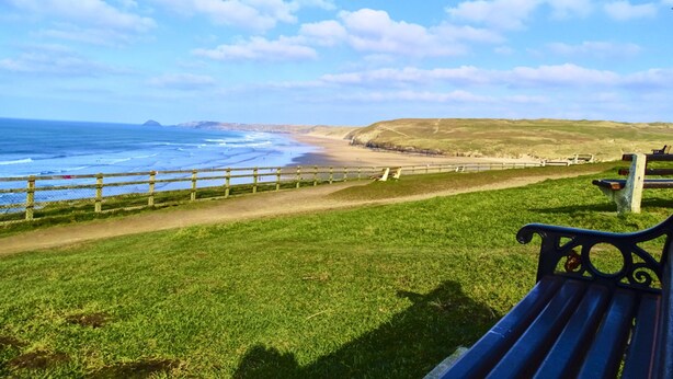 Un espace verdoyant avec un banc, un sentier menant à la plage et une vue sur l’océan et les collines.