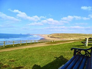A grassy area with a bench, a path leading to the beach, and a view of the ocean and hills. - Tolcarne in Perranporth (Perranporth)