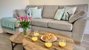 Un salon avec un canapé gris, une table basse en bois, un vase rempli de fleurs roses et une assiette de croissants.