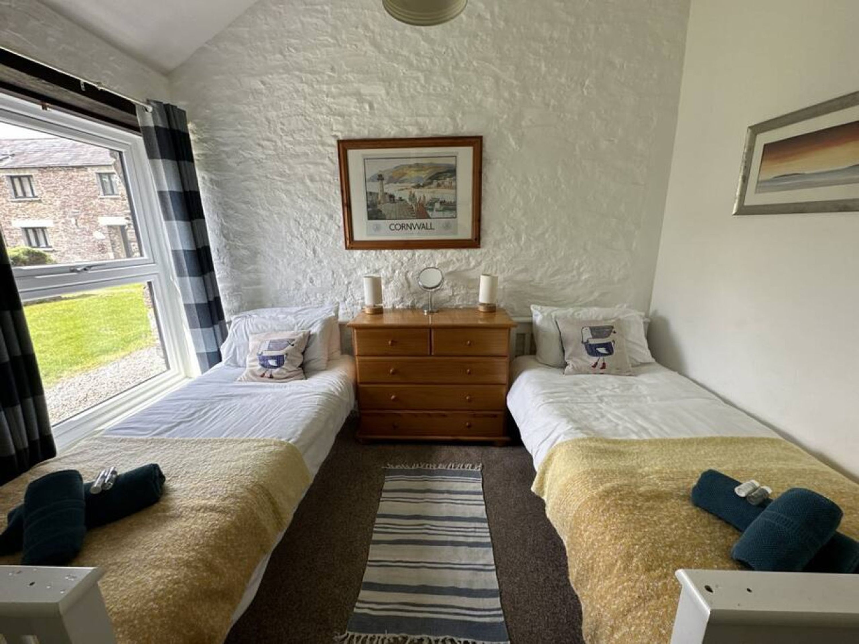 A bedroom with two single beds, a wooden dresser, a framed picture of Cornwall, and a window with curtains.
