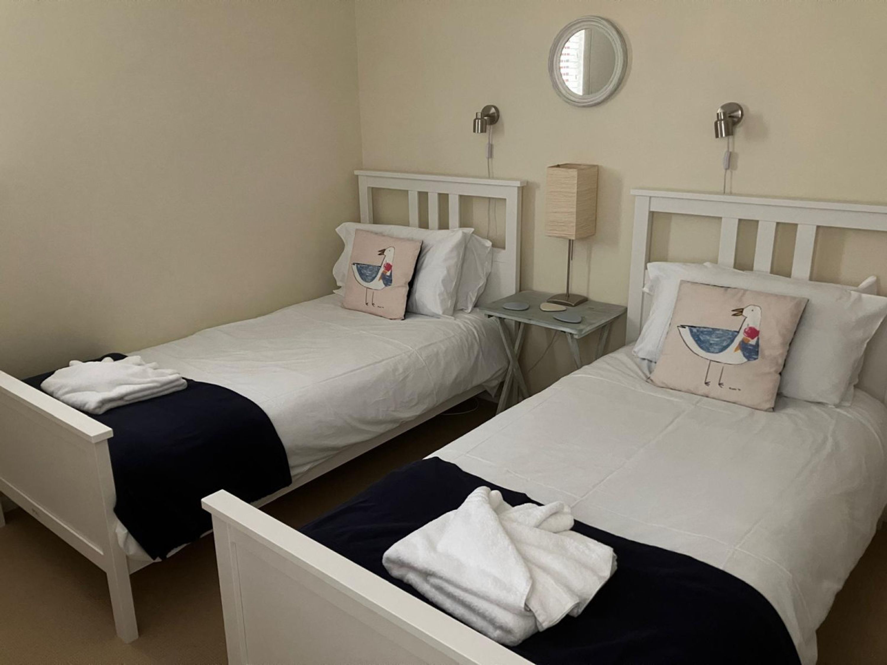 Two single beds with white headboards and matching pillows, a bedside table with a lamp, and a round mirror on the wall.